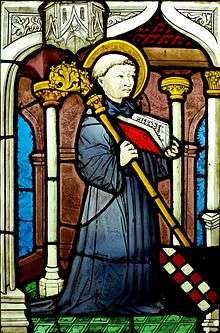 Stained glass image of a kneeling man with a halo holding an open book and a staff.