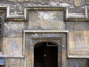 A stone archway, without a door; above, the Roman numerals "VI" in a white circle and, in chalk, two crossed rowing oars with green blades and the words "Blades", "2001", "Men's 1st Torpid"; to the left, "Balliol SEH Keble Merton"; to the right, a standing dragon holding a flag above the initials "JCBC"