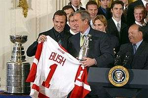 A group of young to older men stand around an older man holding a red and white ice hockey jersey bearing the word "BUSH" and the number "1"