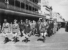 Black and white photo of a large group of middle-aged men marching in close formation down an urban street. The front rank of men are wearing military uniforms, and the remainder are wearing formal suits. Some of the men in suits have medals pinned on their coat lapel.