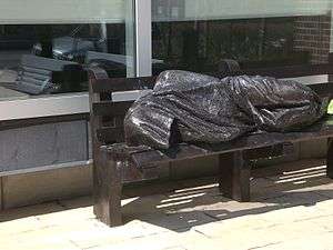 A photograph of a bronze statue of a person covered in a blanket and lying on a park bench all in front of a building with glass windows on a sunny day