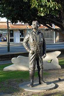 Bronze, life size statue of a man wearing the clothes and equipment of an aviator. A street and buildings can be seen in the background.