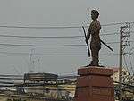 Side on view of a cement statue depicting a man with a moustache holding a rifle in one hand and a stick in another, wearing traditional clothes. He stands on a polished stone pedestal, and a city landscape and buildings can be seen in the background.