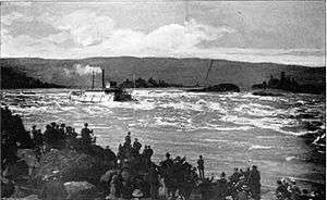 A river boat with more than a dozen windows along its visible side runs a set of rapids on a very large river. Smoke or steam rises from its smokestack and flows behind the boat parallel to the water. In the foreground, a crowd of 50 or people watch the boat from the rocky shore.