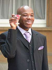 Wiltshire holding his MBE high in his right hand. He is shown from the waist up, smiling and formally dressed (black suit and waistcoate; white shirt with lilac tie, loosely tied). His head is shaved; a ring is visible on his right little finger