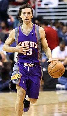 A white basketball player, wearing a purple jersey with a word "Phoenix" and the number "13" written in the front, dribbles the ball