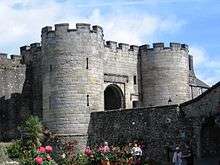 Front of a stone castle gatehouse with round towers in the corners, battlements and an arched entrance.