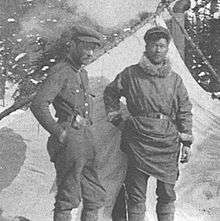 In a grayscale photograph, two men stand in front of a tent and snowy evergreen trees