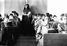 Students in the classroom, with a teacher in nun's garb at the back of the room.