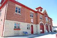 A two-storey concrete and red brick fire hall. There are three large doors for vehicles, one regular door, and two windows.