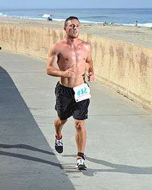 A young man competing in the 2014 Carlsbad Triathlon jogs on a paved path along a beach in Southern California. His expression shows the labor of his effort.