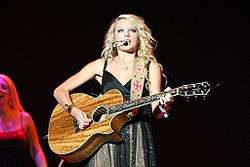 A female teen with blond hair and blue eyes, clothed by a sparkly dress, faces forward and plays a koa wood guitar.
