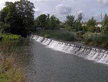 A weir with water flowing from right to left, surrounded by trees and vegetation.