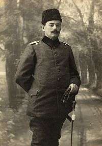 A man in an Ottoman army uniform, sporting a full moustache, and wearing a wool hat and round spectacles. He carries what appear to be gloves in his left hand. His right arm is bent and his right hand rests on the small of his back.