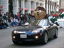 A black open-top sports car with lights on is being driven down an asphalt road. A large furry toy costume, slightly larger than a human is standing in the back seat. It has cream coloured mouth and chest, and dark brown arms and forehead, large whiskers, a grin, large white eyes and two canines. Behind him are some men walking in green costumes. On the left is a crowd watching the parade from the footpath, in front of tall buildings with stone arches.
