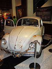 A light tan rusty Volkswagen is positioned for display behind a chain made of handcuffs