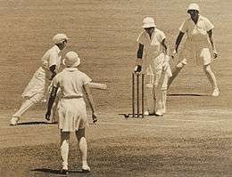 Monochrome image of four women on a cricket pitch, all the women are wearing white knee length sports dresses. The two players right by the stumps are also wearing pads, while the person behind the stumps (wicketkeeper) is also wearing gloves, the woman in front (batsman) is also holding a bat and looking at the stumps. The bowler has her back to the camera while the other woman in the frame is behind the wicketkeeper and facing the stumps.