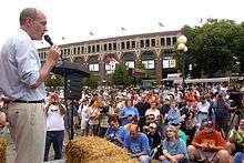 McCotter stands outside with a microphone before a podium and bale of hay facing a seated crowd