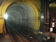 A narrow railway tunnel with a single railway track, lit by a bright white light