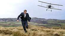 A man in his thirties dressed in a suit being chased across grassy moorland and towards the camera by a biplane.