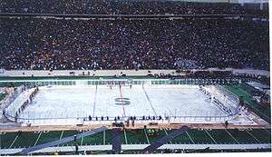 Two teams of many hockey players stand at either end of a rink built on top of a football field as thousands of people look on from the stands.