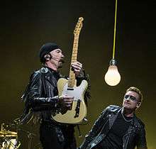 The Edge and Bono clothed in leather jackets, as the The Edge holds a guitar vertically. A large dangling light bulb hangs between them.