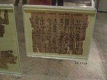 A museum display of an ancient fragment of a papyrus document safeguarded by sealed thick glass, with cursive hieratic handwriting in black ink on its surface