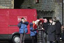 Tennant, Piper, Tate, and Barrowman stand in front of a van that carries the branding of a fictional company in Doctor Who, which is parked outside a church. Tennant is holding a video camera and is filming the "David Tennant's Video Dairies" featurette for the series DVD and Tate is holding an open umbrella coloured in the Welsh national colours.