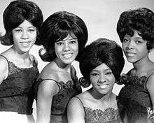A black-and-white photograph of four African-American woman smiling in the direction of a camera. They all have dark hair and are wearing dark-colored tops.
