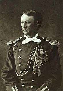 Head and shoulders of a white man with mustache. He is wearing an ornate military jacket with shoulder boards, a scarf around the neck, and cords and tassels hanging from the left breast.