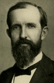 cropped shoulder high portrait wearing a suit and tie, mustache and short beard