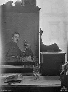 A dresser with a large mirror and a bottle of wine, a glass and some papers sitting on top. A man in military uniform can be seen in the mirror's reflection. He has his hand in his pocket, and is using a camera on a tripod.