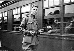 A young man in military uniform holding something in his hands stands beside a train carriage that has men looking out.