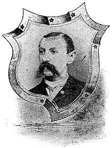 Head of a white man with a drooping mustache and short hair, wearing a dark suit over a light-colored shirt and tie. The portrait is surrounded by a shield-shaped decorative frame.