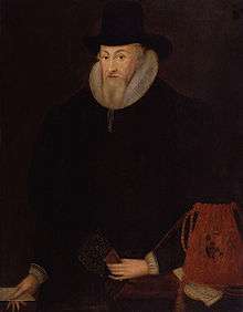 An elderly gentleman dressed in a black coat and hat, with a white ruff around his neck. He has a pointed, grey beard, and is holding a red bag with a coat of arms on it.