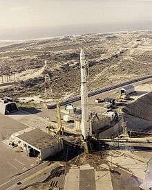 Photograph of the Thor-Agena vehicle on the launch pad at Space Launch Complex 10, surrounded by scrub vegetation and various support buildings, the Pacific Ocean in the distant background.