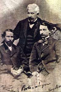 An old photograph depicting two dark-haired men seated in the foreground and a white-haired man standing behind