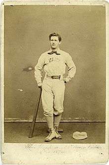 A baseball player is standing, facing the camera, with the top of a baseball bat in his right hand, and the bottom resting on the ground