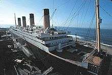 A ship resembling the Titanic is being built at a port with clear skies and small waves.