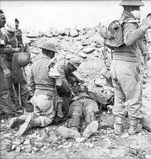 A German doctor and a British soldier kneel over a wounded German soldier who is laying on the ground. Three soldiers, one of whom appears to be wounded, stand to the left while two more soldiers stand to the right.