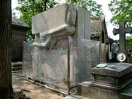 A large rectangular granite tomb. A large, stylised angel leaning forward is carved into the top half of the front. There are a few flowers beside a small plaque at the base. The tomb is surrounded by a protective glass barrier that is covered with graffiti.
