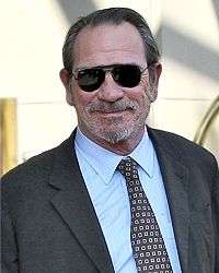Photo of a smiling Caucasian male in his early sixties who is wearing sunglasses, a grey suit, a blue shirt, and a black tie with grey squares and black circles.