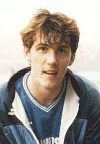 A brown-haired man in a blue football shirt with a blue tracksuit top over it, smiling slightly