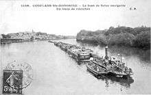 An old black-and-white photograph of a steamboat pulling many barges behind it.