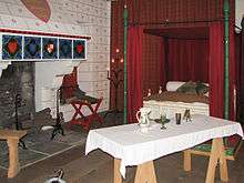 On the left is a fireplace with various heraldic arms painted on it, on the right is a four-post bed, and in the front is a set table on trestles. The floor is wooden and the walls are covered with painted patterns and drapes.