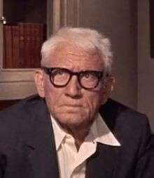 Image of Spencer Tracy with white hair, wearing thick-rimmed glasses and looking grumpy.