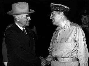 Truman in a dark suit and tie and light hat shakes hands with MacArthur, in uniform wearing a shirt but no tie and his rumpled peaked cap.