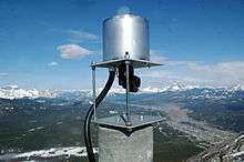 A metallic cylinder sits atop a square platform raised by two bolts. A prism connected to cables monitors the mountain for movement.