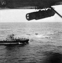 An aerial photograph of a surfaced submarine in choppy sea; a small ship is close by and both appear to be stationary. Part of the aircraft's left wing can be seen, with a bomb hanging from it.