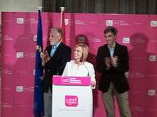 Smiling woman standing at a podium, flanked by two applauding men